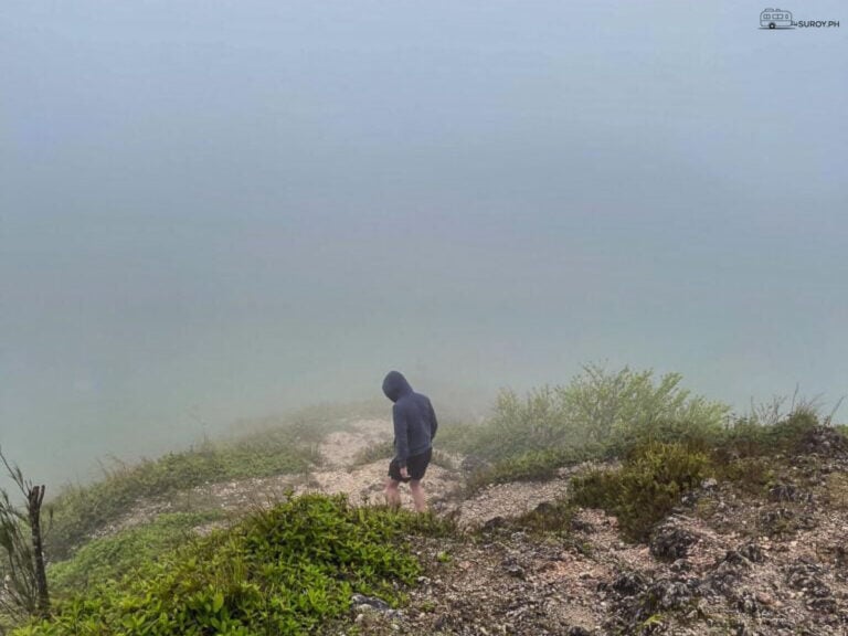 A lone hiker walks a dirt path along the ridge at Osmena Peak in thick fog, low scrub on both sides.