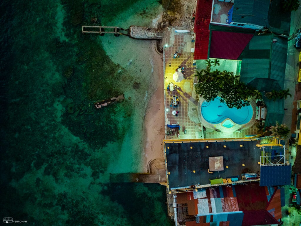 Top down drone frame of the Panagsama waterfront at dusk, a resort pool and tiled terrace on the right, a strip of pale sand, then shallow reef running out into darker water on the left.