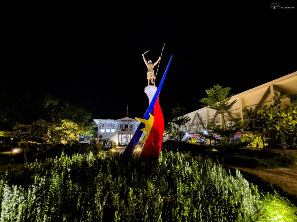 A statue of a child fighting for freedom on top of the Philippine Flag with Museo Naga in the background. 