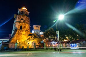 The coral stone campanario in Dumaguete lit warm at night, market stalls and a street light flare beside it.