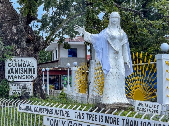 A serene statue standing near Guimbal Vanishing Mansion, with signs highlighting the local legend and conservation efforts.