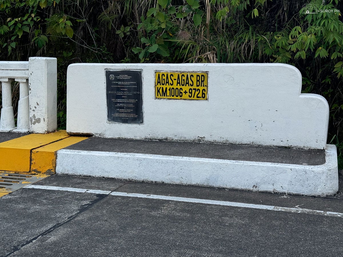 The bridge nameplate on the concrete parapet, marked AGAS-AGAS BR. KM 1006 plus 972.6 in black on yellow.