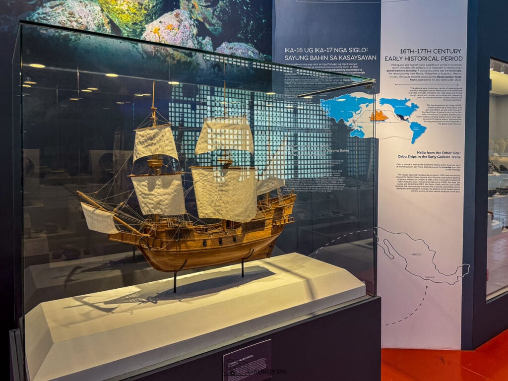 A wooden model galleon in a glass case beside a wall map of the galleon route