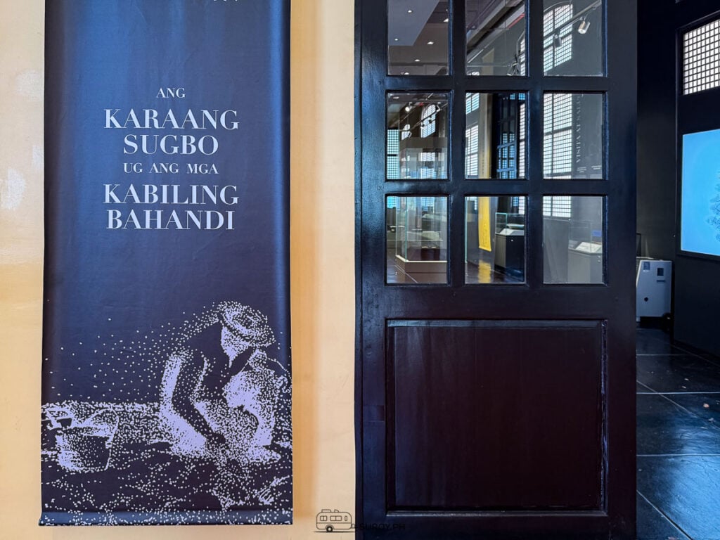 A tall exhibit banner reading Ang Karaang Sugbo ug ang mga Kabiling Bahandi beside a dark wooden door