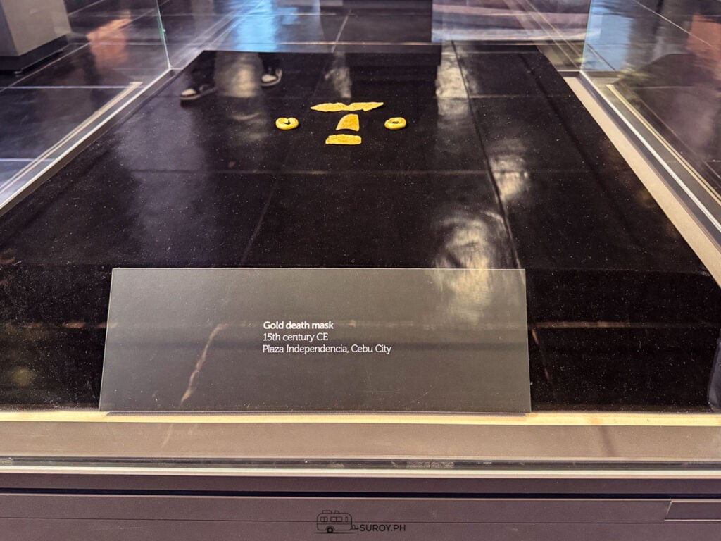 The gold death mask in a low glass case, small gold pieces for the eyes, nose and mouth on a black surface, with its label