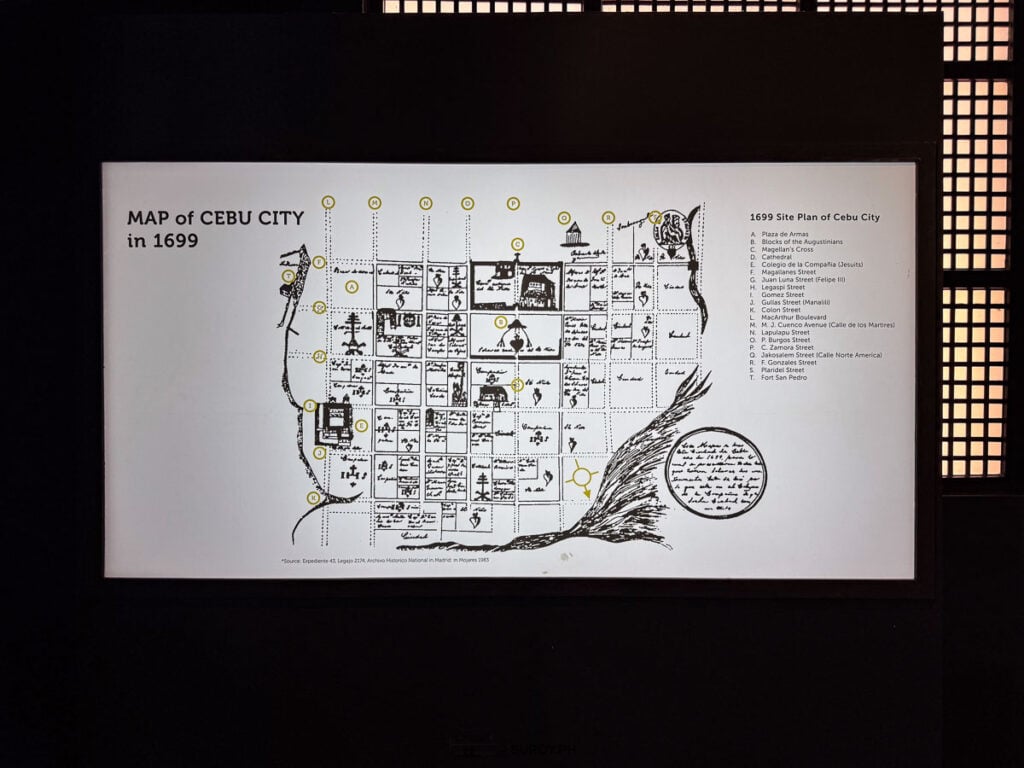 A lit museum panel showing a hand-drawn map of Cebu City in 1699 with lettered points and a key