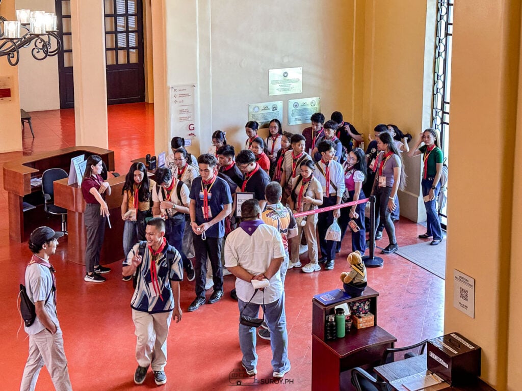 A group of students in uniform lining up in the red-floored entrance hall