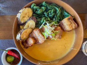 A beautifully plated serving of pork belly kare-kare with a creamy peanut sauce, fresh greens, and eggplant crisps—a must-try dish at Kaayoo.