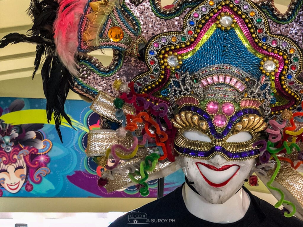 A MassKara mask in close up, beaded in blue, pink and gold with a wide painted smile and feathers rising behind it.