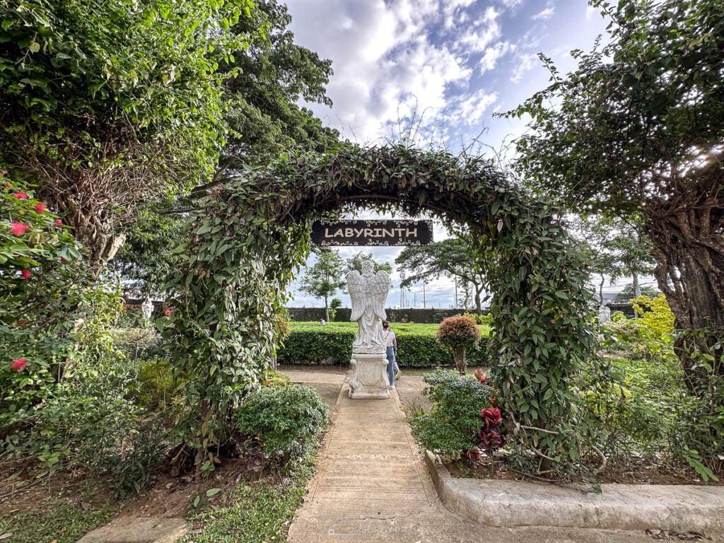 An inviting entrance to the labyrinth garden, framed by lush greenery and crowned with a wooden archway, promising a path of calm and introspection.