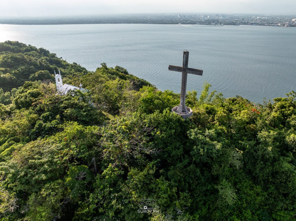Aerial view of Sinukuan Hill in Guimaras Island with a large cross, forested slopes, and ocean coastline in the background