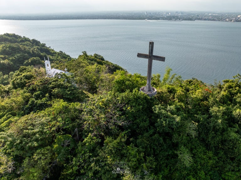 Aerial view of Sinukuan Hill in Guimaras Island with a large cross, forested slopes, and ocean coastline in the background