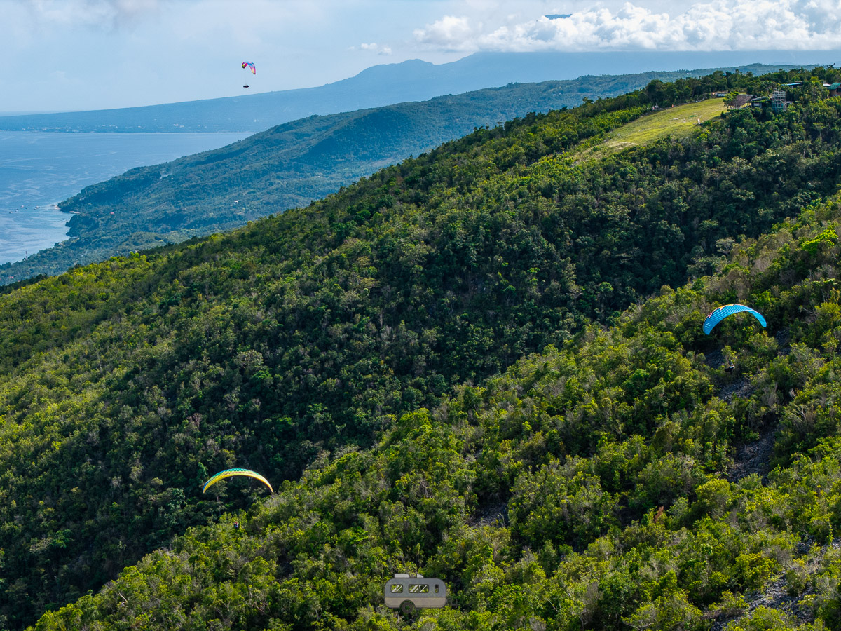 Three paragliders in the sky over a lush green mountain ridge in Oslob, facing the sea.
