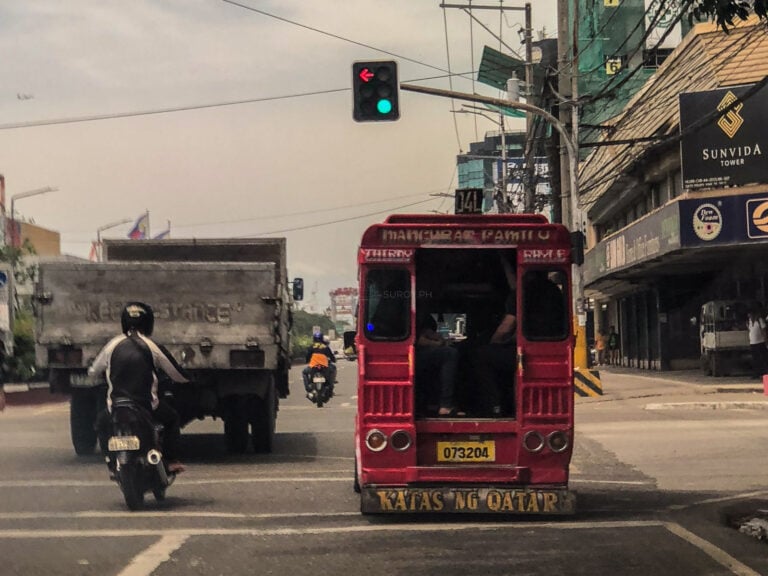 The view from behind on a Cebu road, a helmeted motorcyclist riding between a dump truck and a red jeepney, shopfronts along the roadside.
