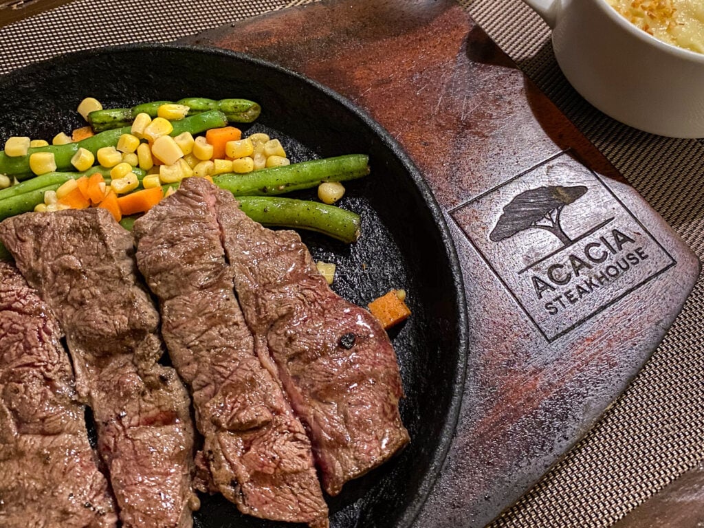 Sliced steak with sweetcorn and green beans on a sizzling cast iron plate set in a branded Acacia Steakhouse wooden board
