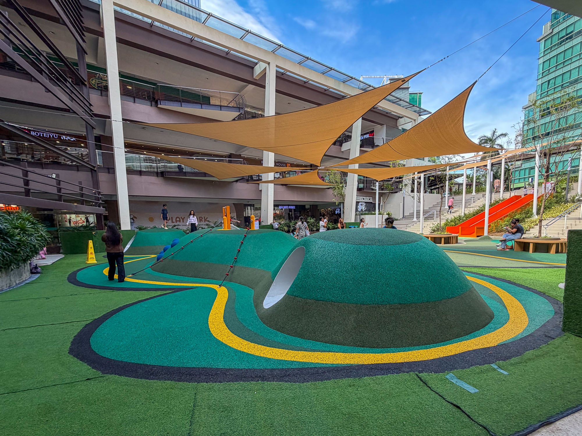 The open-air play park at Ayala Center Cebu: rubberized green hills and tunnels under shade sails, with an orange slide on the terraced steps behind.