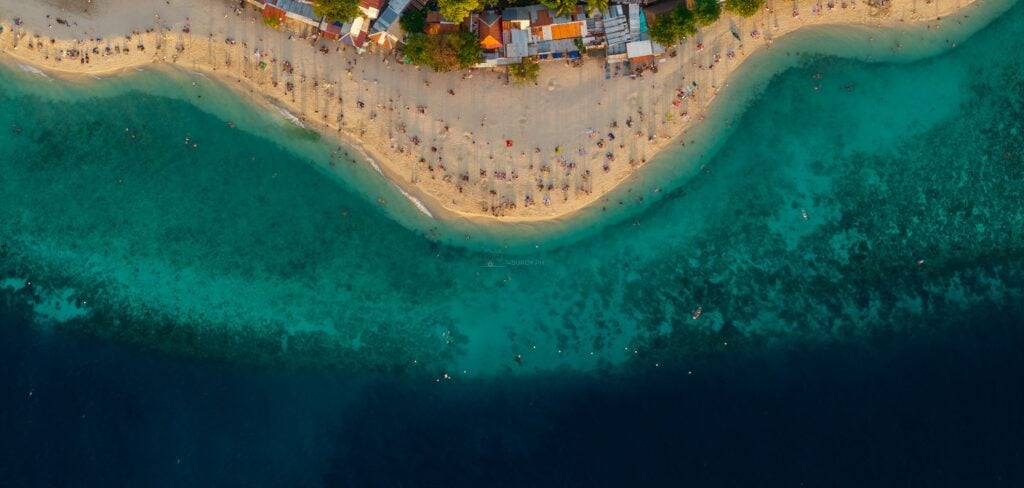 Aerial view straight down over Basdaku, the white beach at Moalboal, Cebu, a curved sand strip lined with umbrellas, pale reef flat below it, and the deep navy of the drop off across the bottom.
