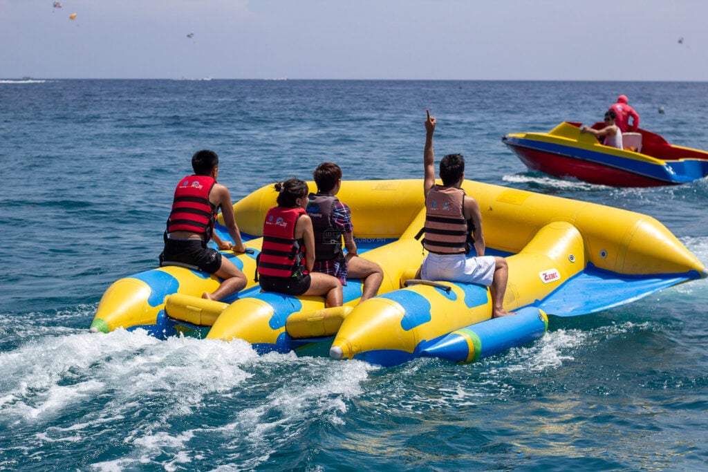 Riders in life vests on a towed inflatable, one raising a hand, with a speedboat alongside off Boracay