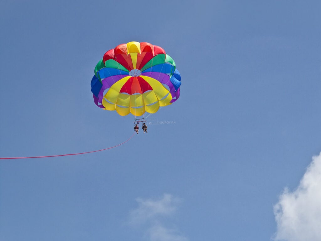 A rainbow parasail canopy carrying two riders high above the sea off Boracay, its tow line running down out of frame