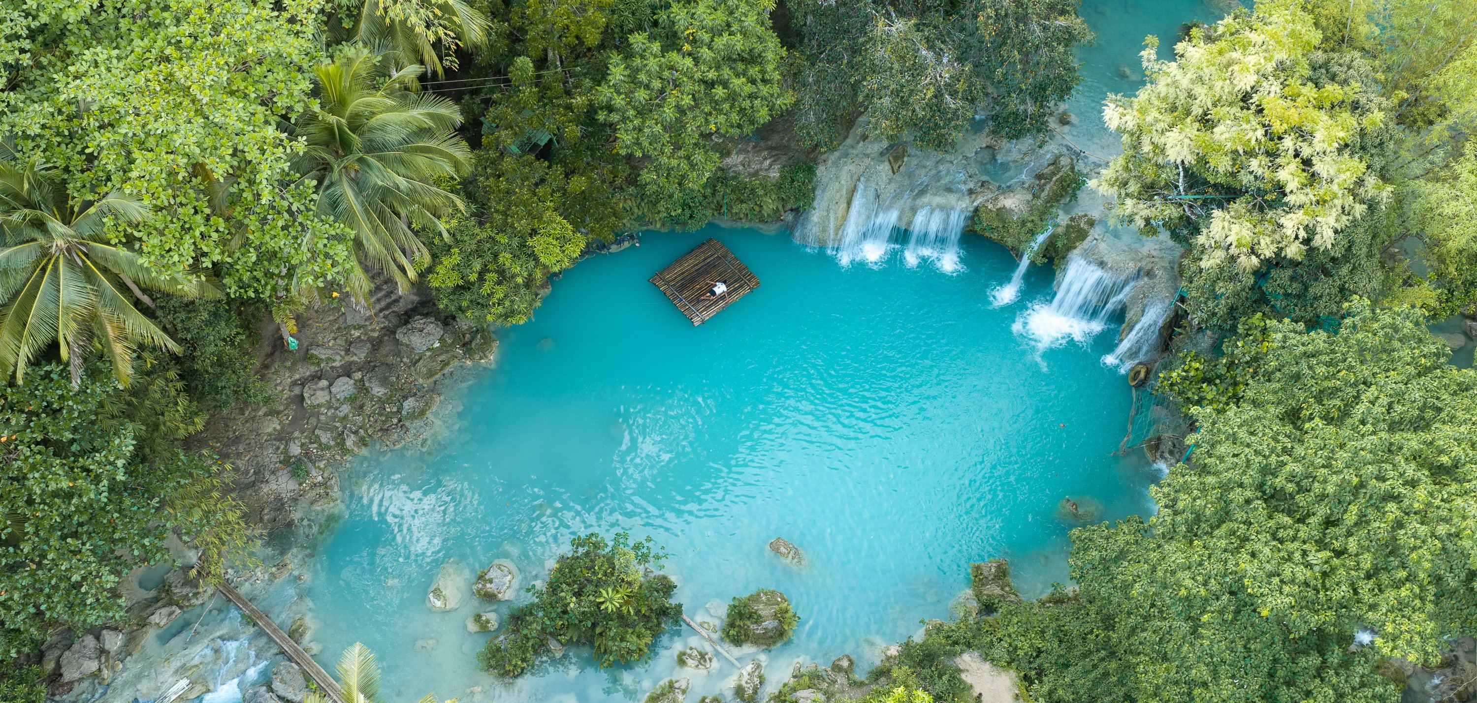 Aerial view of Cambugahay Falls in Lazi, Siquijor, water stepping down three tiers into a turquoise pool with a bamboo raft floating on it