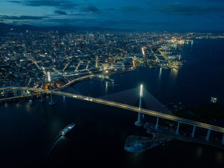 The full span of the Cebu Cordova Link Expressway at blue hour, both pylons lit, a passenger boat crossing the channel below and the Cebu City skyline and harbour stretching behind it.