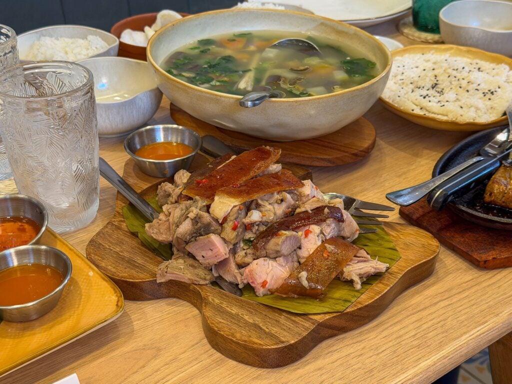 Table with carved lechon on a wooden board lined with banana leaf beside a bowl of soup and plates of rice