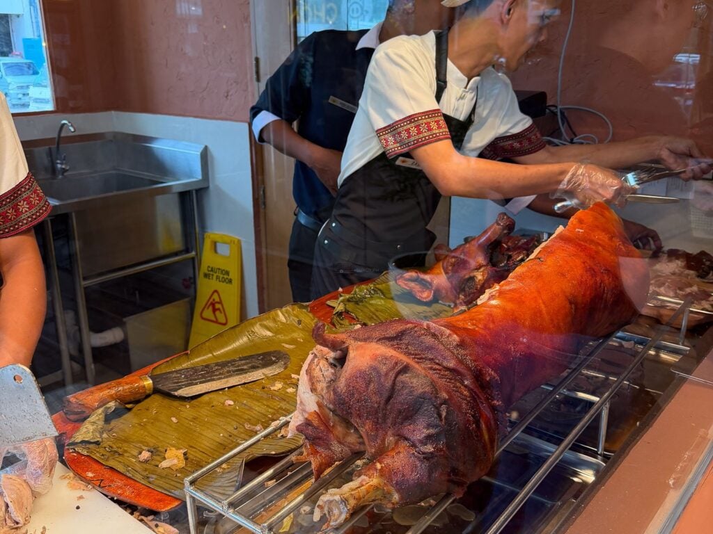 Cook in an embroidered apron carving a whole roast pig on a rotisserie behind glass at a Cebu lechon house