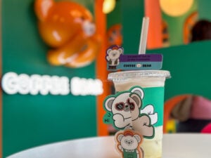 An iced layered Coffee Bear drink in a clear cup with a bear character sticker, against the green wall of the Cebu IT Park branch
