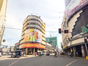 View down Colon Street with a curved corner building carrying fried chicken signage, jeepneys parked on the right and pedestrians crossing