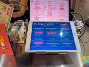 Tablet on a stainless steel cafe counter displaying a blue filter coffee menu listing origins, varieties and roaster with a pink printed food menu behind it