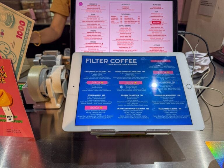 Tablet on a stainless steel cafe counter displaying a blue filter coffee menu listing origins, varieties and roaster with a pink printed food menu behind it