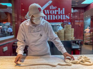 A Din Tai Fung cook in a white coat and cap rolling dumpling dough at the counter in front of the red world famous wall at SM City Cebu