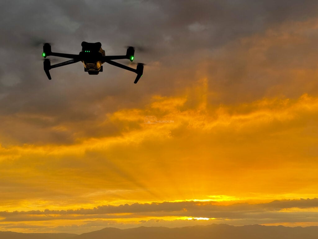 A drone hovering high in the frame against an orange and gold sunset with mountains on the horizon below