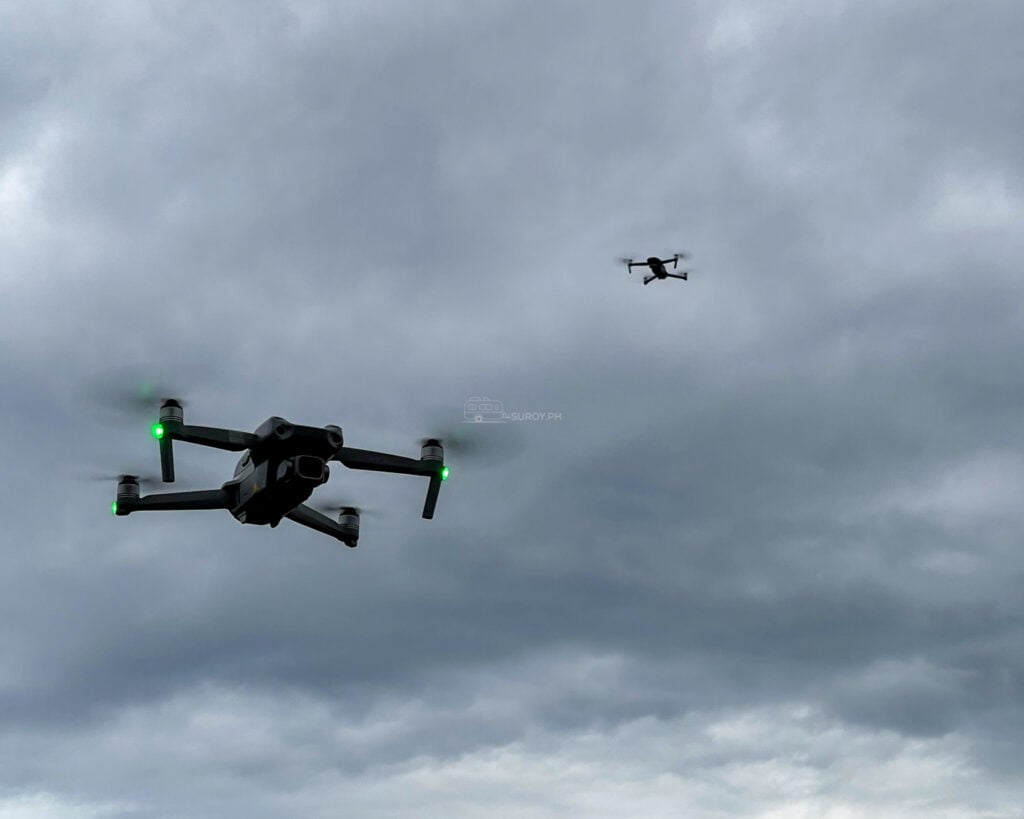 Two drones flying under a heavy grey overcast, the nearer one a DJI Air 2 with the model name visible on its arm and the second already a dot in the distance