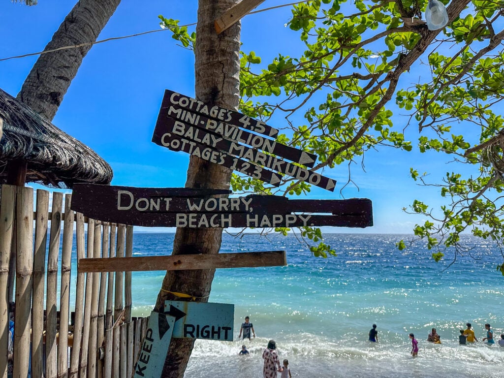 Wooden signpost on a Guimbal beach pointing to cottages and a mini pavilion, swimmers in turquoise water behind it