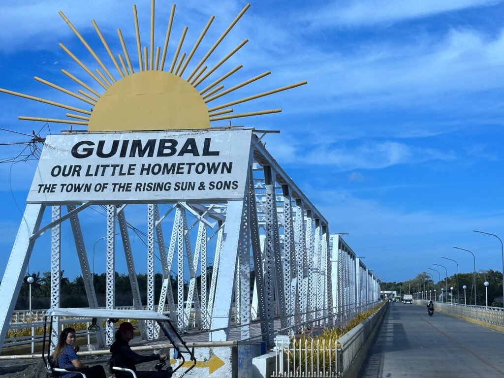 Steel truss bridge in Guimbal, Iloilo beside a sign reading Guimbal, Our Little Hometown, The Town of the Rising Sun and Sons