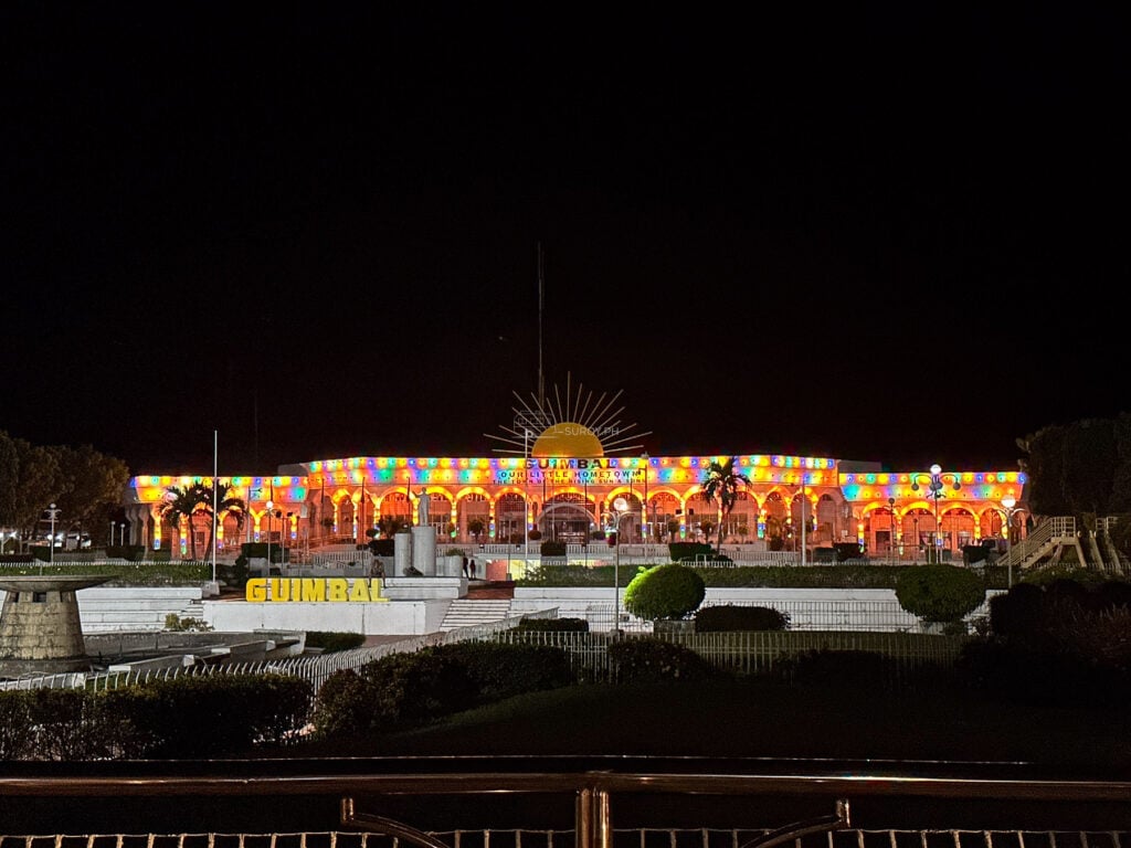 Arcaded civic building in Guimbal, Iloilo lit in color at night, the town name in lit letters under a rising sun emblem