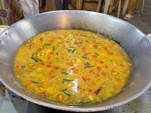 Overhead view of a large steel wok filled with golden fish broth, halved yellow and orange tomatoes and whole green chillies