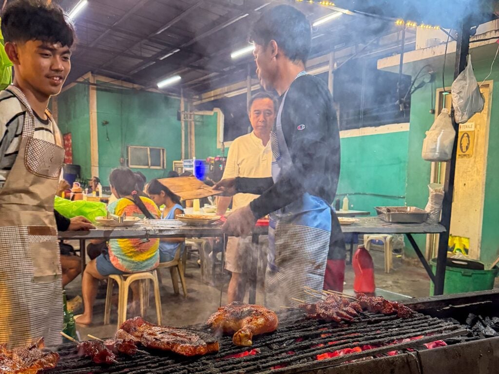 Cooks tending pork and chicken barbecue over a charcoal grill with diners seated at long tables behind