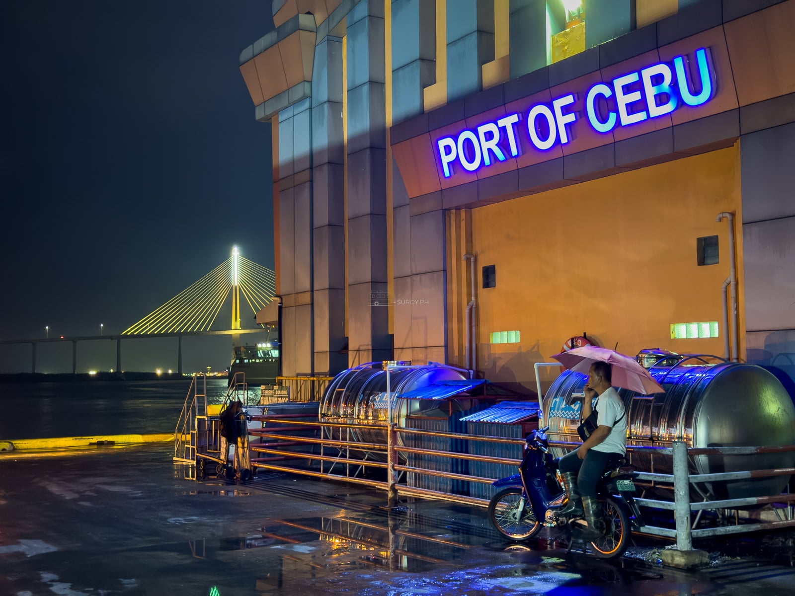 The lit Port of Cebu sign on the passenger terminal at night in the rain, with the CCLEX bridge glowing across the channel and a motorbike rider waiting under an umbrella