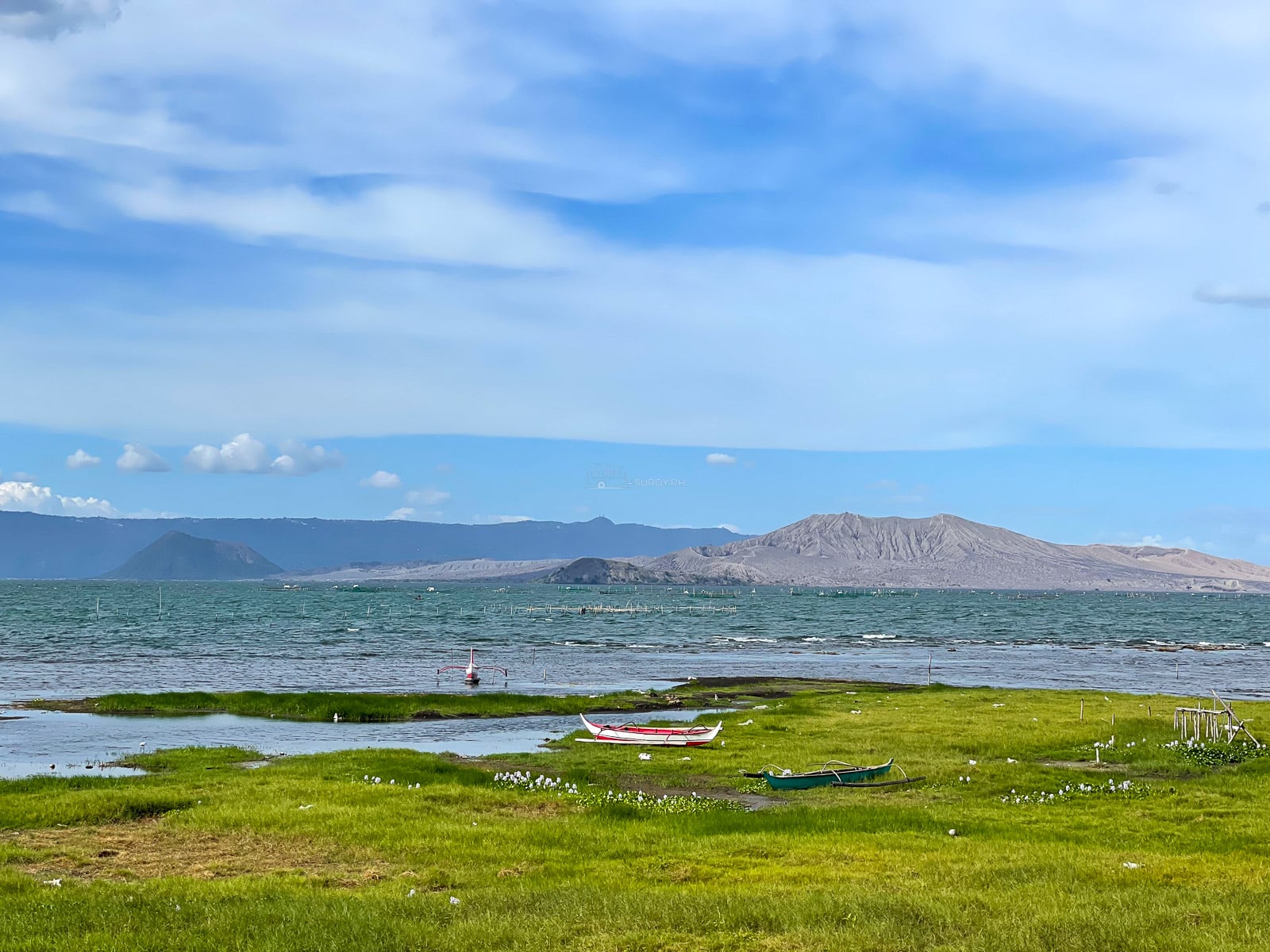 Grassy shoreline of Taal Lake with a small red boat and the volcano across the water