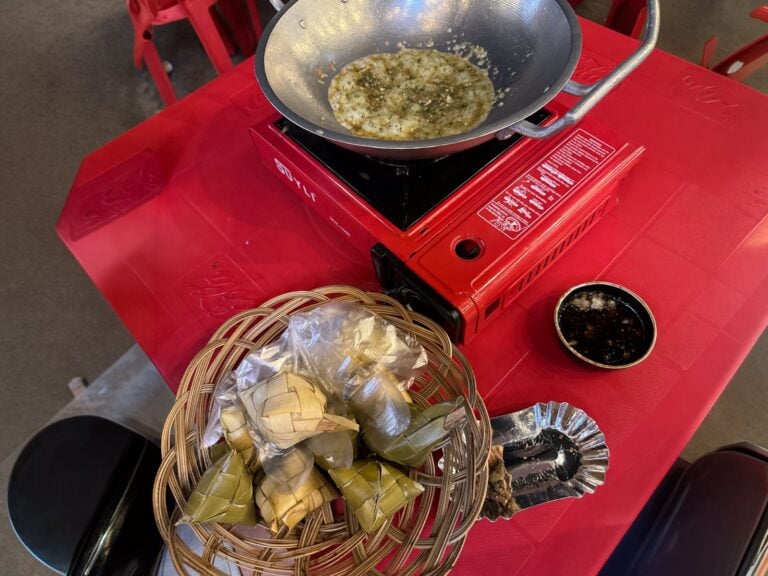 Overhead view of a wok of bubbling tuslob buwa on a red portable stove beside a rattan basket of woven rice pouches