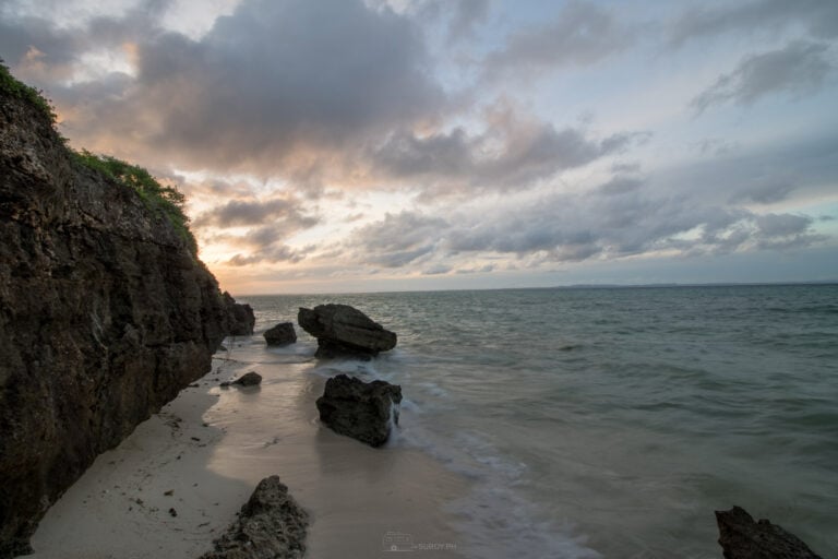 Low cliff and scattered boulders on the Bantayan Island shore at 5:47 in the morning, the water smoothed by a long exposure under heavy cloud
