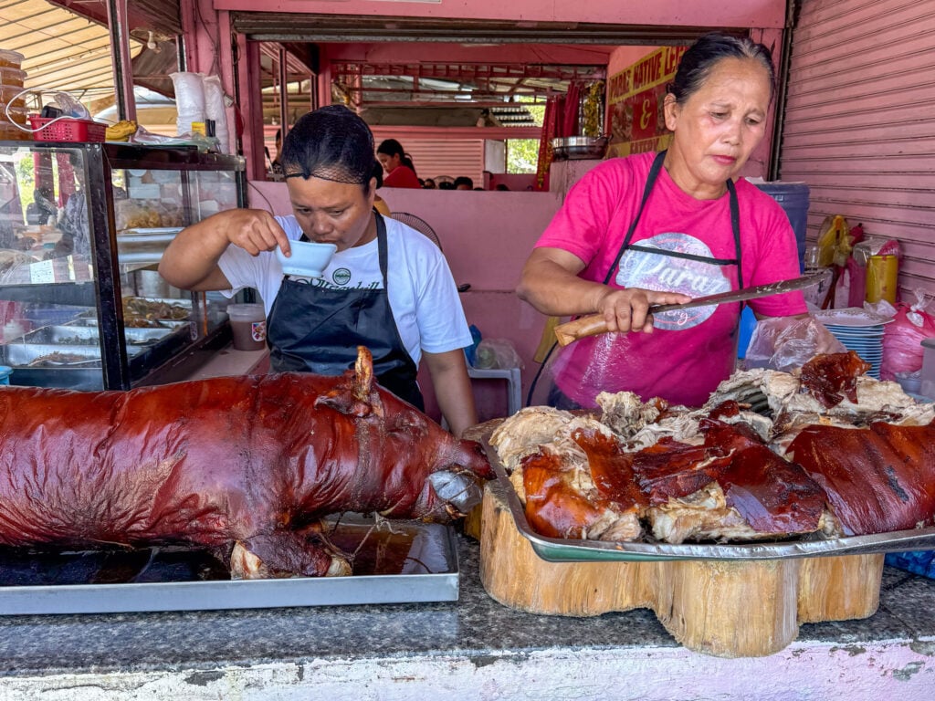Two vendors working behind a Carcar lechon counter with a whole roast pig laid out in front of them