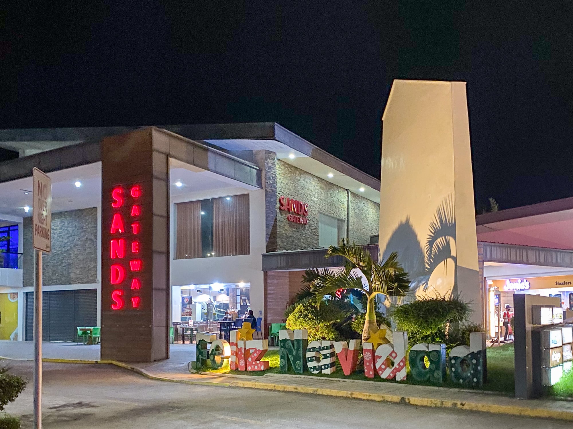 Sands Gateway complex in Danao City lit up at night with a Feliz Navidad letter display and palms out front
