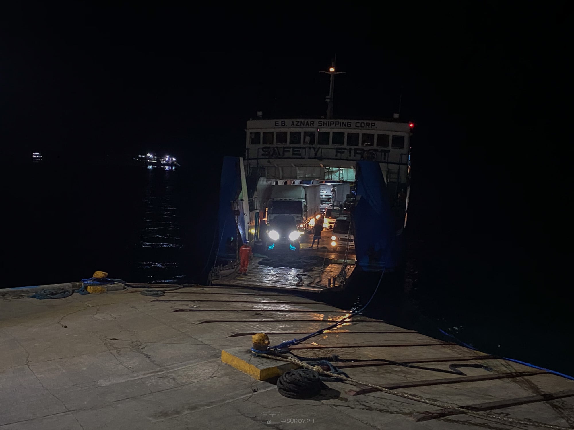Roll-on roll-off ferry moored at Danao pier at night with its bow ramp down and vehicle headlights inside the car deck