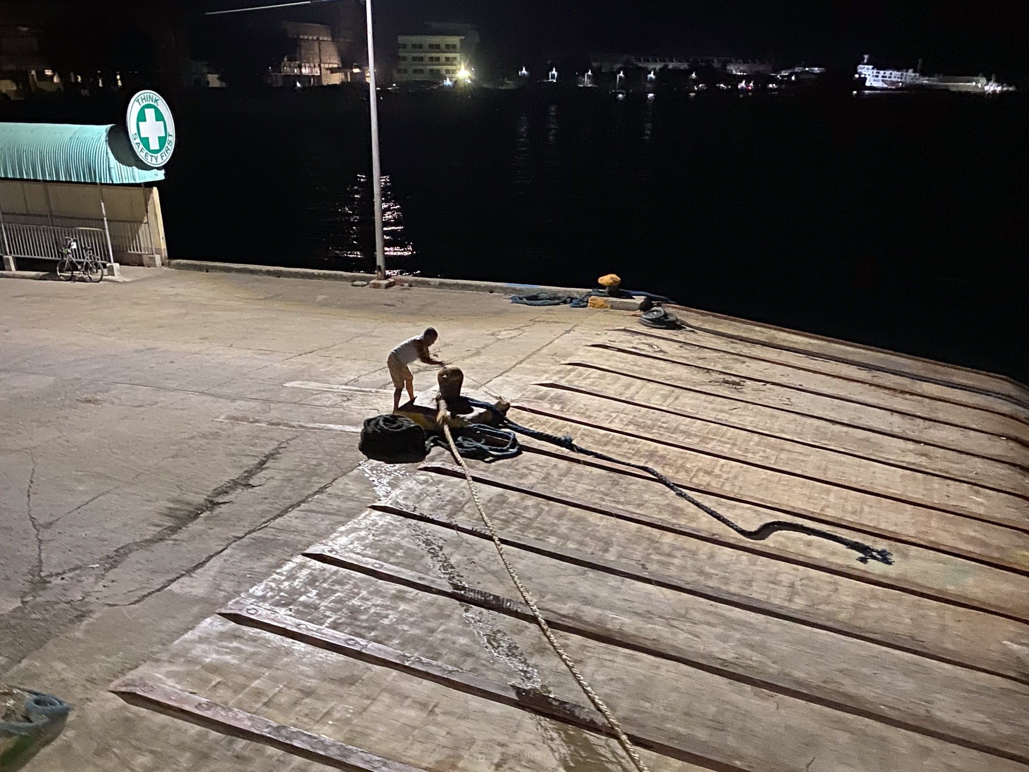 Crew member coiling mooring lines on the concrete pier apron at Danao port at night, with the lights of the town across the water