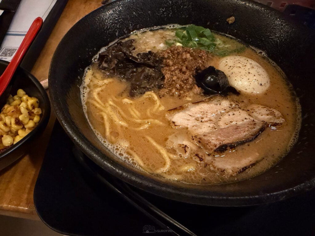 A black bowl of paitan ramen at Hamakaze with chashu, half an egg, minced dark topping, wood ear mushroom and spring onion, with a side bowl of butter corn beside it