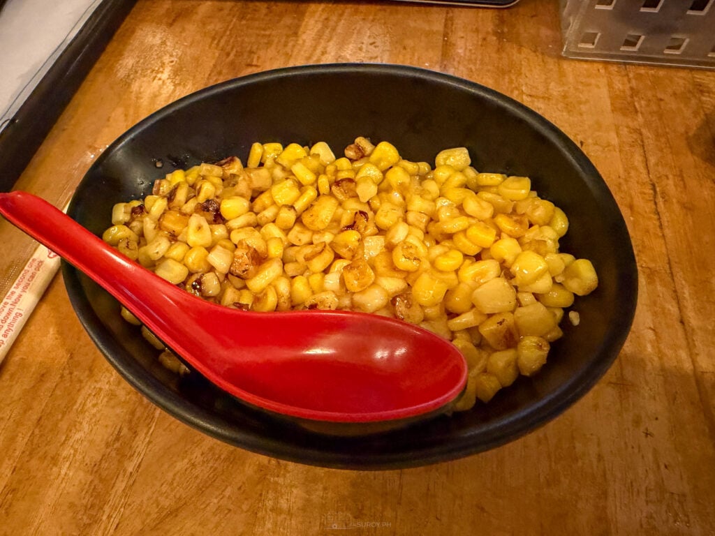 A black bowl of butter corn at Hamakaze Ramen, kernels browned at the edges with a red spoon resting in them on a wooden table