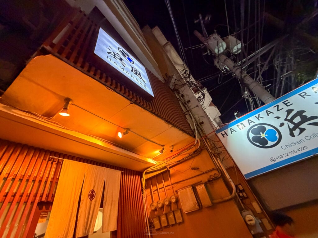 The lit exterior of Hamakaze Ramen on A.S. Fortuna at night, with a blue sign reading Hamakaze Ramen Chicken Collagen and a warm orange lit entrance behind a noren curtain